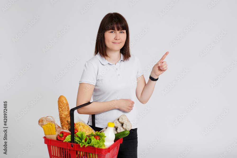 portrait of young woman with shopping basket full of products pointing at something over white portrait of young woman with shopping basket full of products pointing at something over white