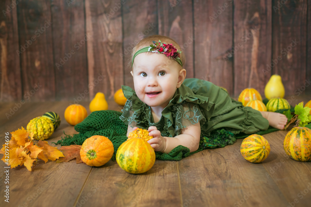 Fototapeta premium Cute little girl up to 1 year old in a green cute dress and a floral wreath with small yellow and orange pumpkins on a brown wooden background