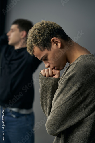 Staged photo illustrates problems and conflicts in gay couple relationships. Moment of showdown: swarthy brooding and upset man is standing in front of his partner in the background. © RedUmbrella&Donkey