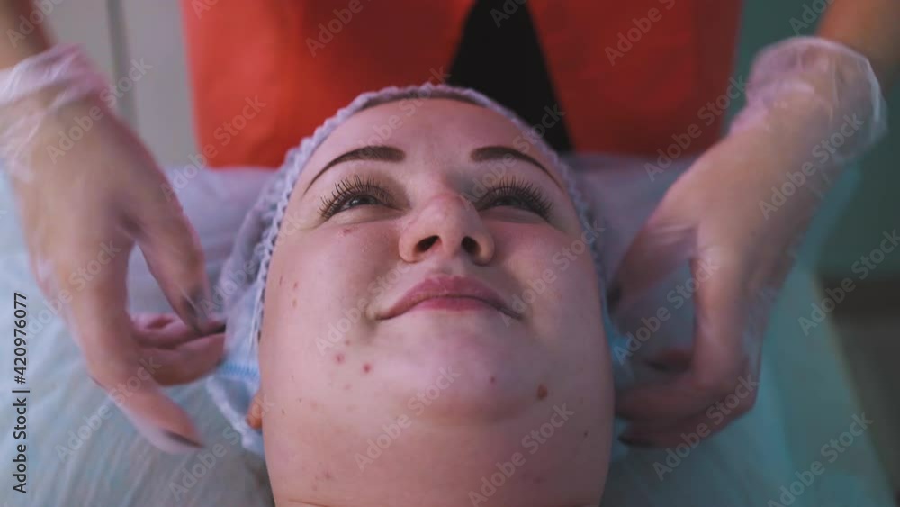 CU: female master in transparent gloves puts protective cap onto beautiful large woman client head before cosmetological procedure in clinic closeup CU: female master in transparent gloves puts protective cap onto beautiful large woman client head before cosmetological procedure in clinic closeup