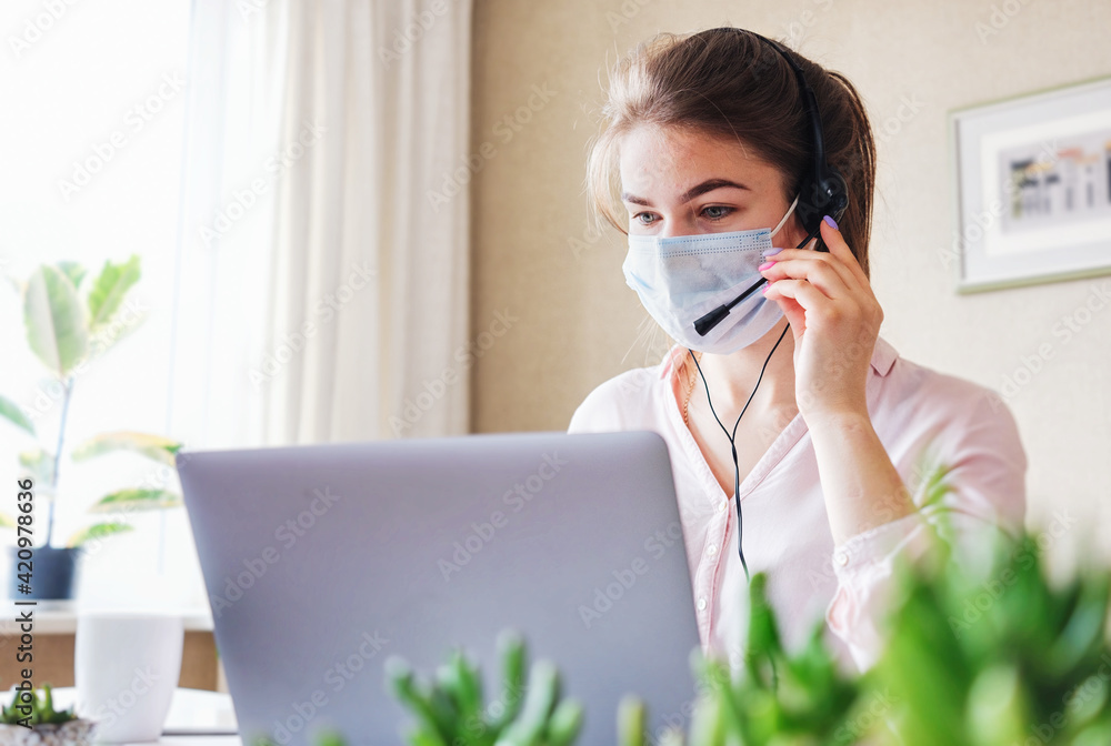 A telephone sales agent wears a headset during the coronavirus pandemic - Hot line operator girl wearing a face mask works in a customer support office - A girl in a call center A telephone sales agent wears a headset during the coronavirus pandemic - Hot line operator girl wearing a face mask works in a customer support office - A girl in a call center