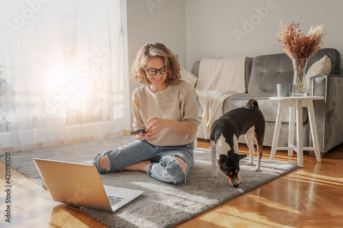 Obraz na płótnie Pretty curly happy young woman sitting at home on the floor in front of laptop with her pet dog working and learning online w salonie