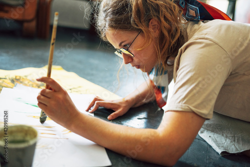 Side view of a female artist sitting on the floor in the art studio and painting on paper with a paintbrush. A woman painter with glasses painting with watercolors in her workshop.