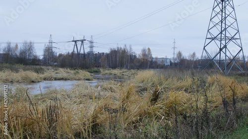 Electric lines in the field with small river and waving grass. Cloudy autumn in Russia