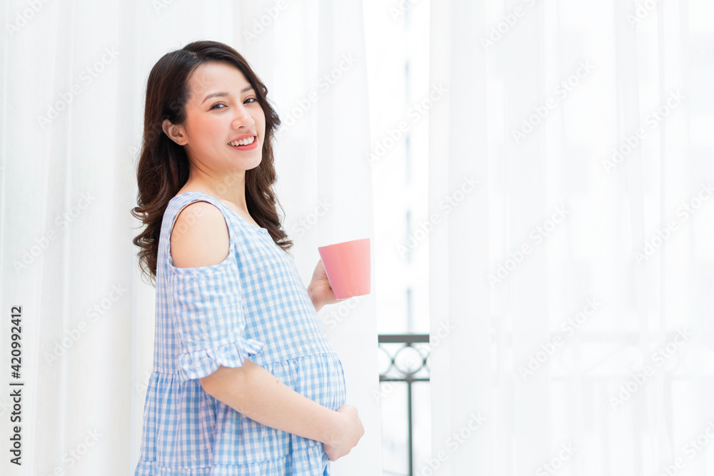 Beautiful woman holding her stomach and holding a cup of milk stands by the window Beautiful woman holding her stomach and holding a cup of milk stands by the window