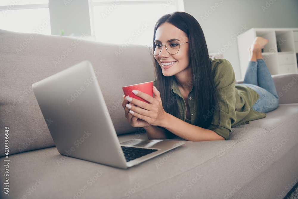 Portrait of lovely cheerful girl lying on divan using laptop watching video pastime drinking cacao at home house flat indoor Portrait of lovely cheerful girl lying on divan using laptop watching video pastime drinking cacao at home house flat indoor