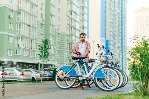 Fototapeta Naklejka Na Ścianę i Meble -  Tourist with a beard and casual clothes takes a sharing bike on the background of the cityscape, enjoys the program on the smartphone. The man rents a bicycle for a walk around the town