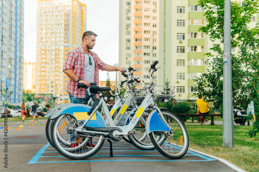 An adult man in a shirt rents a bicycle for a walk on the weekend. City bike rental via smartphone, man puts the bike in place after riding. City online bicycle rental.