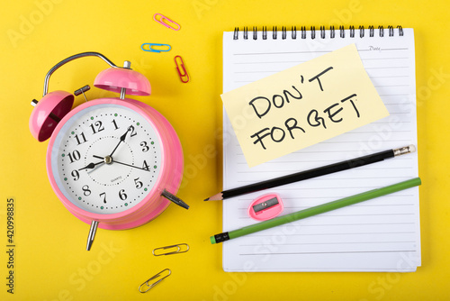A table clock with Don't forget words Written on a sticky note with notepad pencil and other elements on a Yellow background.