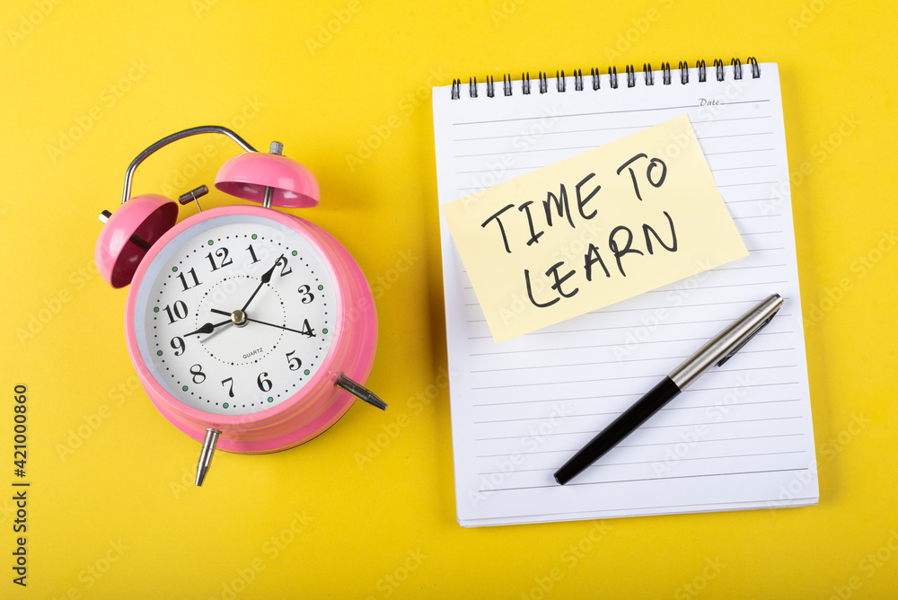 A table clock with Time to learn words Written on a sticky note with notepad and other elements on a Yellow color background A table clock with Time to learn words Written on a sticky note with notepad and other elements on a Yellow color background