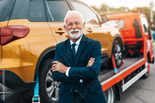 Elegant senior businesman is happy and satisfied with fast and reliable towing service for help on the road. He is standing in front of wrecker with arms crossed. © hedgehog94
