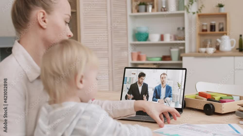 Side view of blurred young Caucasian woman sitting by desk at home, holding blond-haired toddler, who playing with her business documents. Mother having online conference with male colleagues Side view of blurred young Caucasian woman sitting by desk at home, holding blond-haired toddler, who playing with her business documents. Mother having online conference with male colleagues