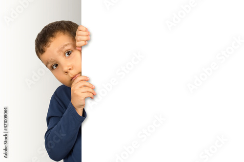 Foto Serious asian boy hiding and peeking carefully behind a blank white billboard