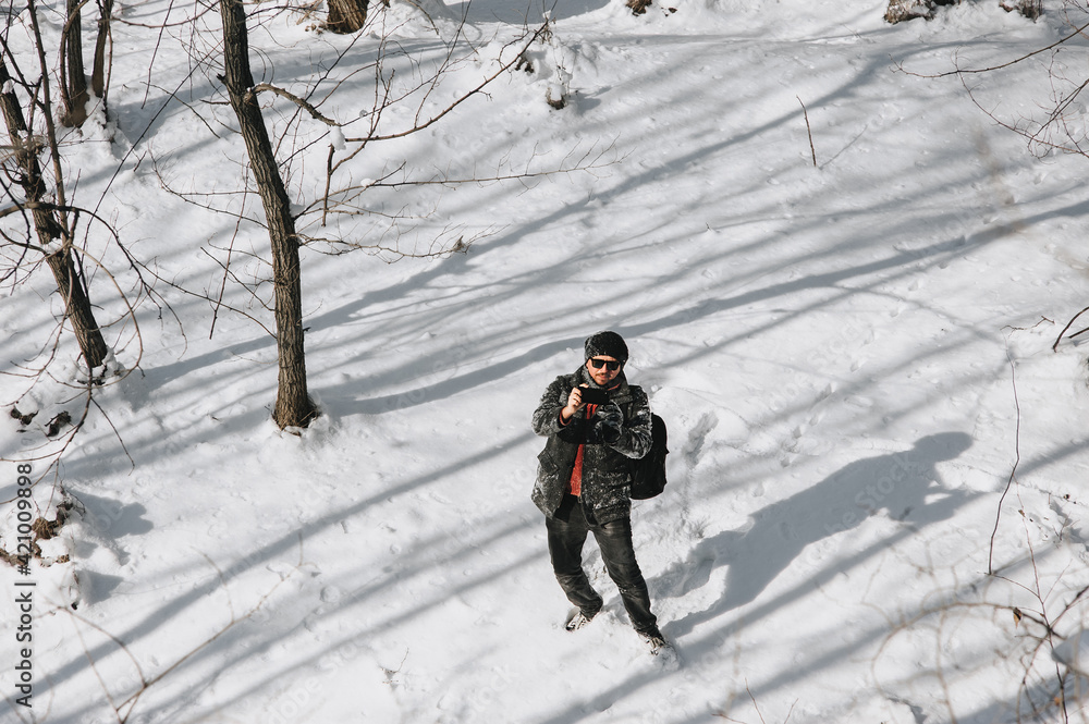 A young man, a wanderer, a tourist in black warm clothes and a large backpack on his back travels through the snowy forest and takes pictures of nature on a mobile phone.