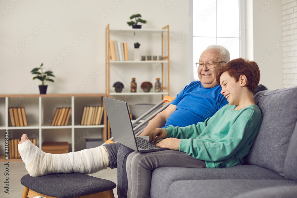 Senior man with injury spends recovery period at home with family. Happy grandson and his grandfather with broken leg in plaster cast sitting on sofa and watching funny movie on laptop computer Senior man with injury spends recovery period at home with family. Happy grandson and his grandfather with broken leg in plaster cast sitting on sofa and watching funny movie on laptop computer