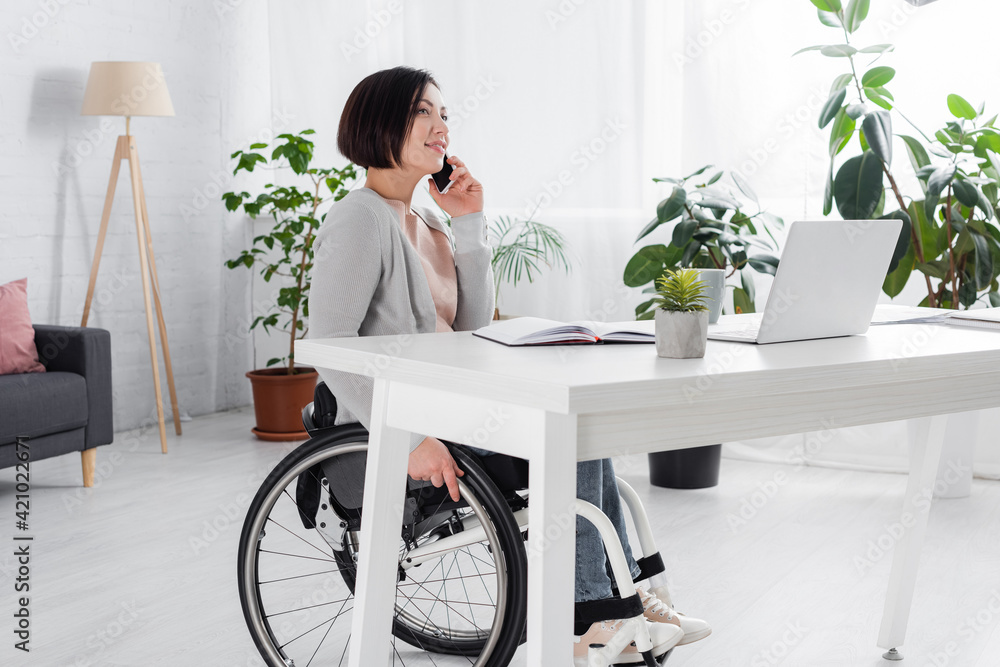 Smiling handicapped woman talking on smartphone near laptop and notebook at home