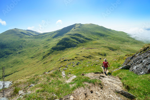 A female hiker and their dog walking down from the mountain summit of Meall Corranaich with Ben Lawers and Beinn Ghlas in the distance in the Scottish Highlands, UK landscape.