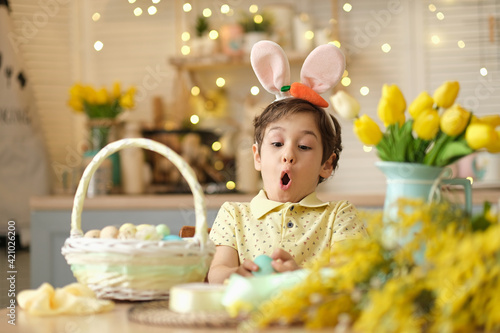 child boy wearing bunny ears sitting in the kitchen at the table. child holding painted eggs preparing for easter. funny surprised child