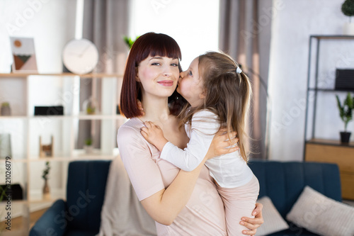 Lovely family mom and child. Happy affectionate young mother, holds her cute smiling little daughter, which is kissing mom in cheek with love, spending free time at home, posing in cozy living room