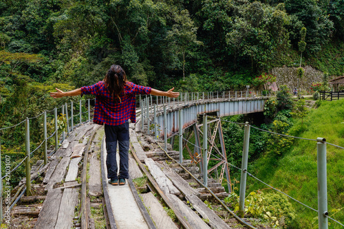Man from behind with dreadlocks and square shirt standing on the train tracks on a bridge with open arms and looking at the sky
