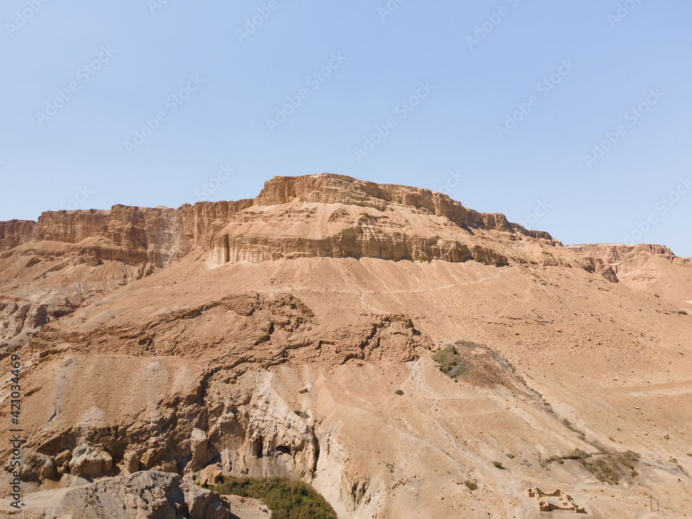 A sandy  mountain that used to be bottom of Dead Sea stands near Ein Bokek embankment on the coast of the Dead Sea, the sea itself and the mountains of Jordan visible in the distance, in Israel