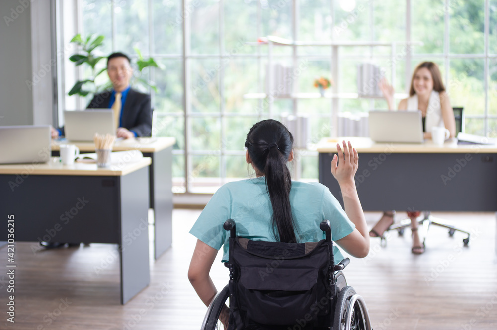 Behind an Asian female office worker in a wheelchair raises her hand to greet a colleague, Disabled work with their colleagues at office workplace Behind an Asian female office worker in a wheelchair raises her hand to greet a colleague, Disabled work with their colleagues at office workplace