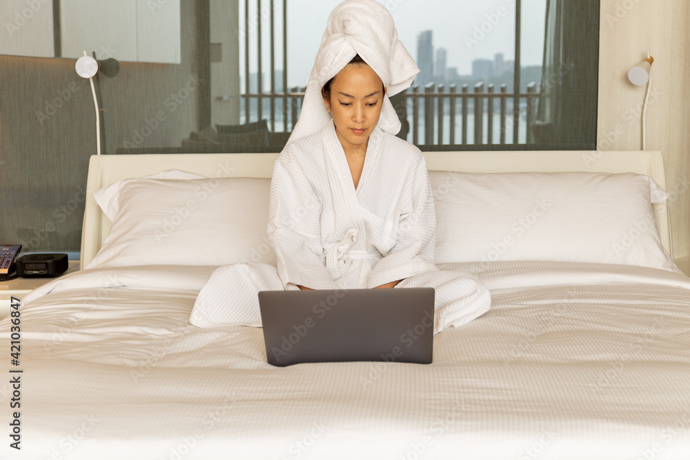Woman in dressing gown and towel on her head sits in bedroom on bed working on laptop. Woman in dressing gown and towel on her head sits in bedroom on bed working on laptop.