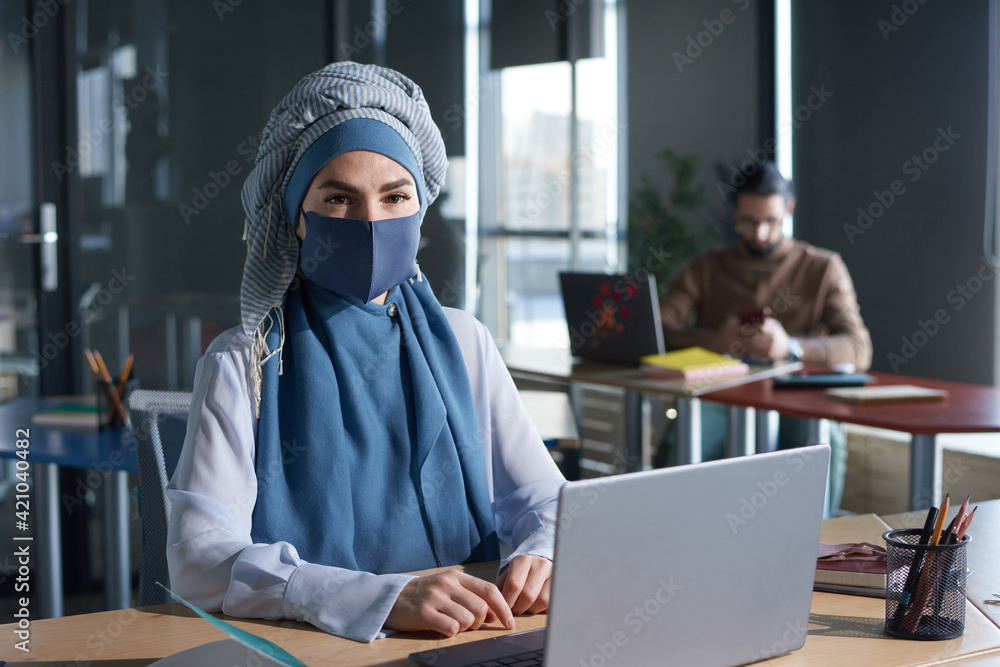 Portrait of young muslim woman in hijab looking at camera while working online on laptop at office