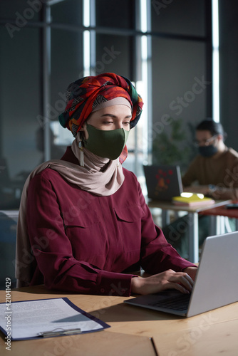 Young businesswoman in hijab and protective mask using laptop for online work during pandemic