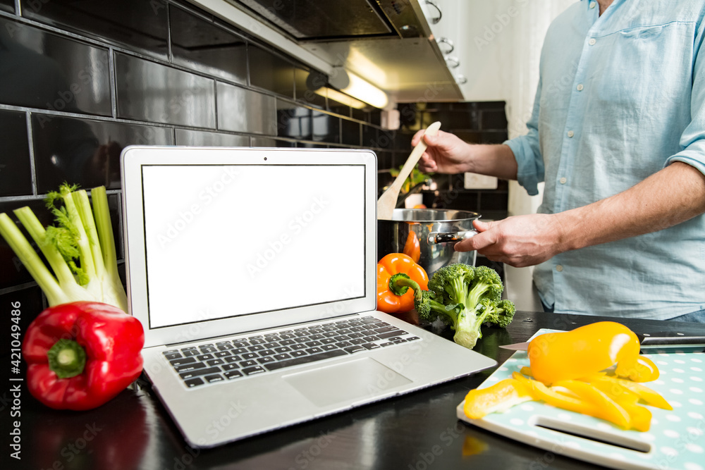 Man using laptop and cooking in kitchen at home. Distant work. Fresh vegetables on kitchen counter. Healthy lifestyle and social media concept. White empty screen Man using laptop and cooking in kitchen at home. Distant work. Fresh vegetables on kitchen counter. Healthy lifestyle and social media concept. White empty screen