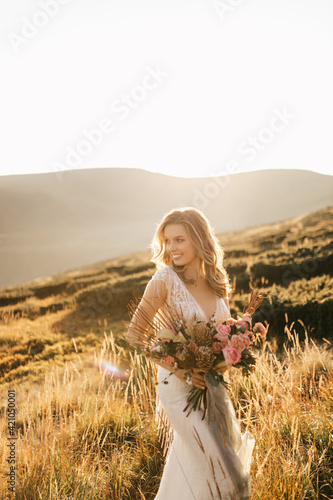 Beautiful bride in a white wedding dress stands posing and smiles in the mountains with a bouquet