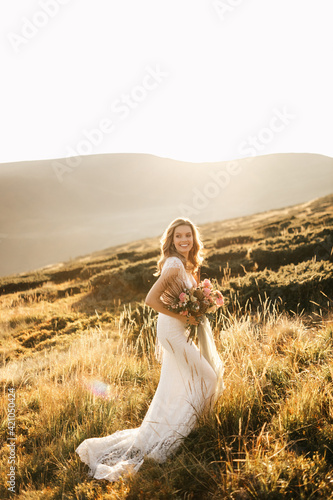 Beautiful bride in a white wedding dress stands posing and smiles in the mountains with a bouquet