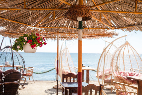 Fototapeta Naklejka Na Ścianę i Meble -  cafe by the sea, restaurant on the beach, thatched beach umbrellas,flowers in pots, table, wicker swing chair, chairs and rocking chair, Crimean pier, views of the mountains and the sea