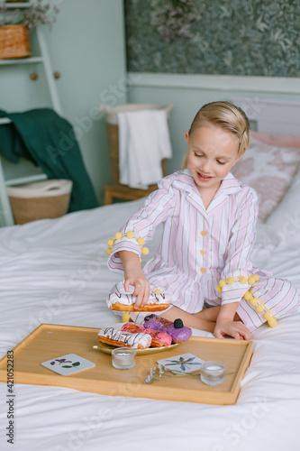 Child sits on the bed in the bedroom next to a tray with sweets Stylish interior Blonde girl with blue eyes in pajamas Breakfast in bed