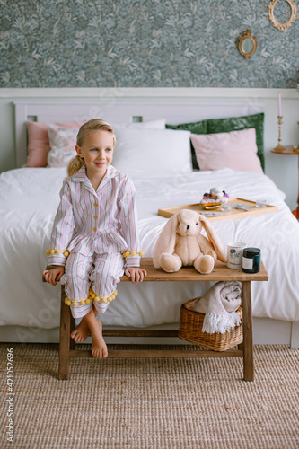 Child girl sitting in pajamas in the bedroom Blonde with blue eyes next to a toy beige plush hare a cup with cocoa and a candle Stylish interior Model portrait