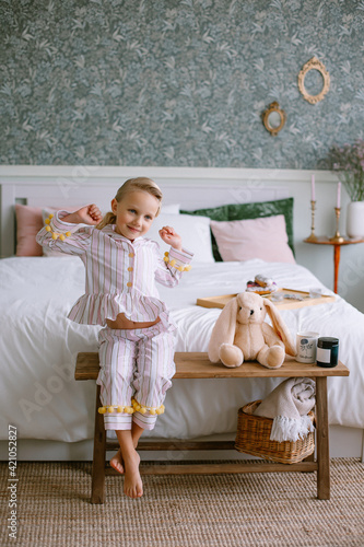 Child girl sitting in pajamas in the bedroom Blonde with blue eyes next to a toy beige plush hare a cup with cocoa and a candle Stylish interior Model portrait