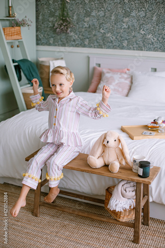 Child girl sitting in pajamas in the bedroom Blonde with blue eyes next to a toy beige plush hare a cup with cocoa and a candle Stylish interior Model portrait