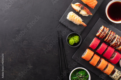 Assorted sushi roll set served on a black slate board, seaweed salad, soy sauce in a bowl and wasabi on the side. Asian food composition on black background, top view, copy space