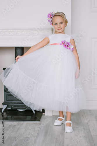 Little girl model in a beautiful white dress outfit White walls and a black fireplace Gray floor Accessory on the head blonde Blue eyes Interior minimalism