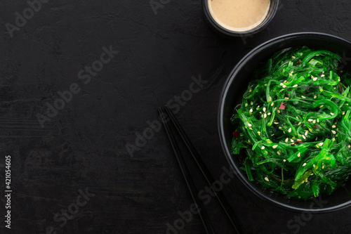 Healthy green seaweed salad with sesame seeds in a black bowl, peanut sauce and chopsticks on a slate black background. Top view, copy space.
