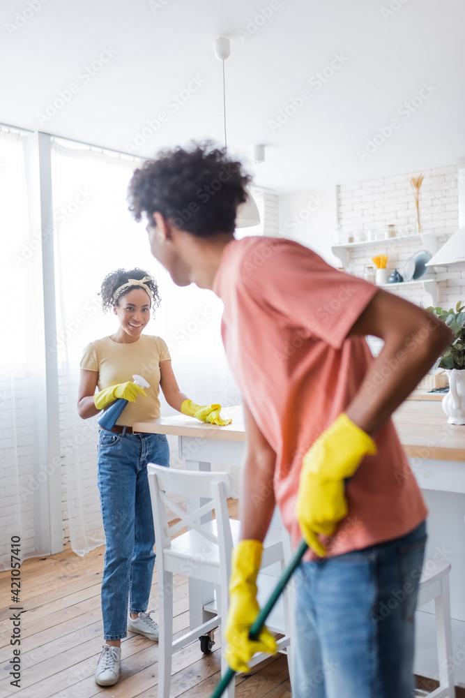 Smiling african american woman cleaning kitchen table near boyfriend with mop Smiling african american woman cleaning kitchen table near boyfriend with mop
