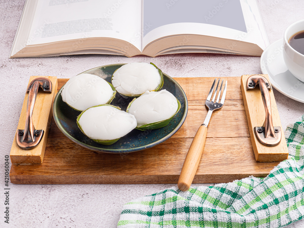 Top view of Thai pudding with coconut topping on a plate on a wooden tray with a book, a white coffee cup, and a cloth placed on a gray stone background. Space for text. Concept of relaxation