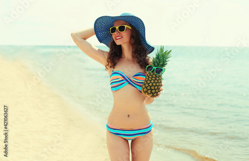 Portrait of happy young woman on a beach with funny pineapple wearing a straw hat on a sea background