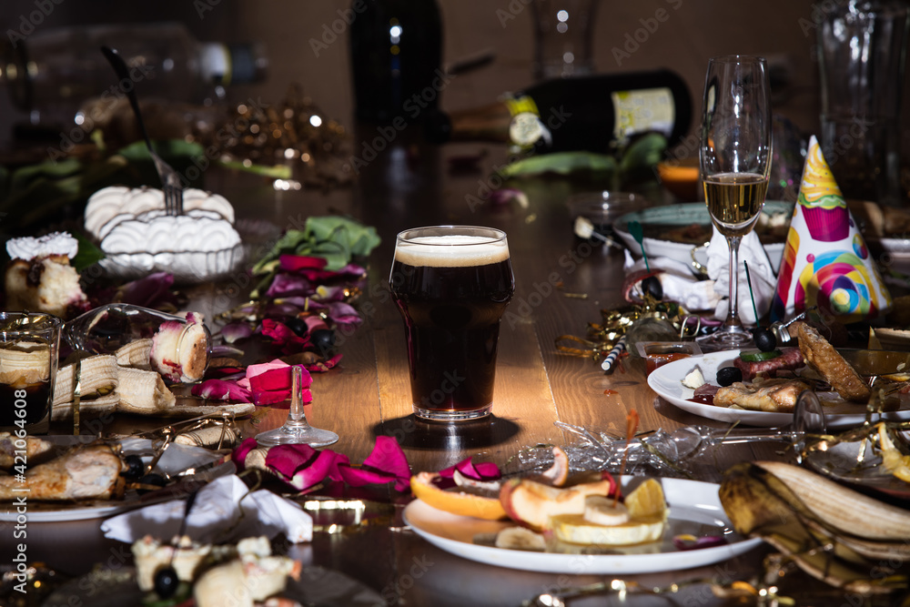 Early morning after the party. Glass of dark, cold stout, beer on the table with confetti and serpentine, leftovers, flower petals