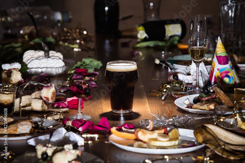 Early morning after the party. Glass of dark, cold stout, beer on the table with confetti and serpentine, leftovers, flower petals