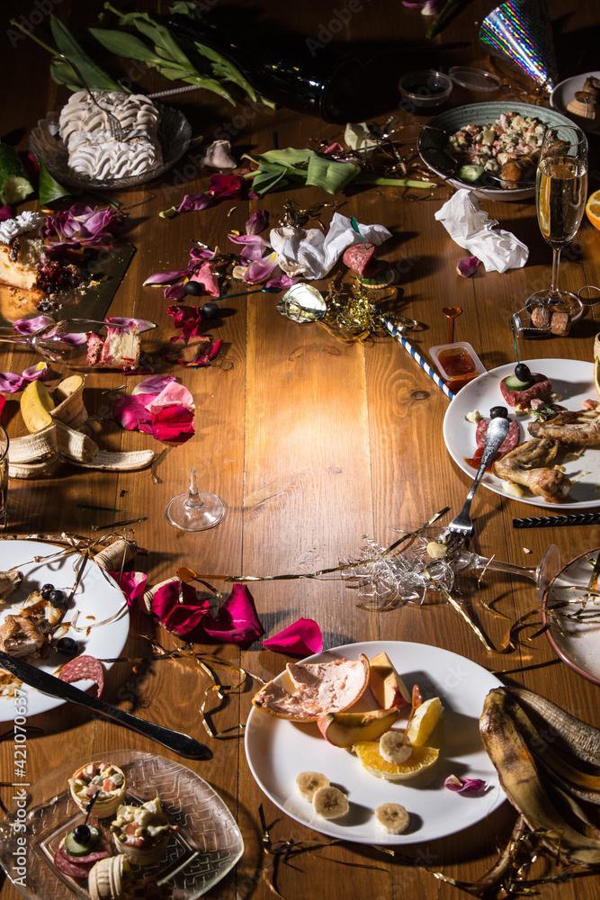 Early morning after the party. Glasses and plates on the table with confetti and serpentine, leftovers, flower petals