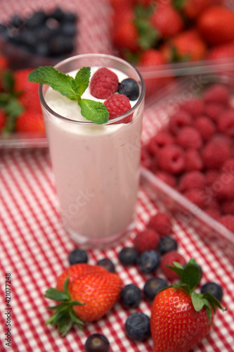 Yoghurt decorated with mint, raspberries and blueberry, on a red and white checkered cloth. With container of raspberries in the background
