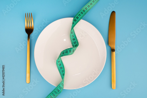 white round plate and golden fork and knife with green measuring tape on blue background, Concept of anorexia and diet