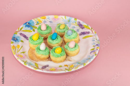 Easter treats made with seven vanilla cookie cups, green frosting nests, and candy eggs on a pastel colored plate against a pink background.