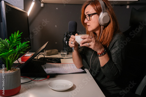 Mujer caucásica de mediana edad tomando un cafe mientras esta grabando un podcast en el despacho de su casa  photo